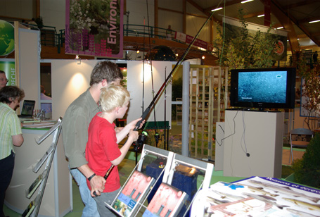 Un membre de la Maison de la pêche luxembourgeoise aide un enfant à pêche grâce au simulateur de pêche